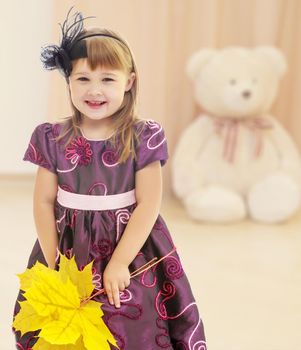 Nice little girl dressed in brown dress. She is holding a bouquet of maple leaves.In the children's room where sits a large Teddy bear .