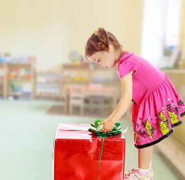 Cute little girl in a pink dress, turned sideways and bent over a large box with a gift. The girl unties the bow.On blurred background the great hall of the kindergarten, with long racks where there are toys.