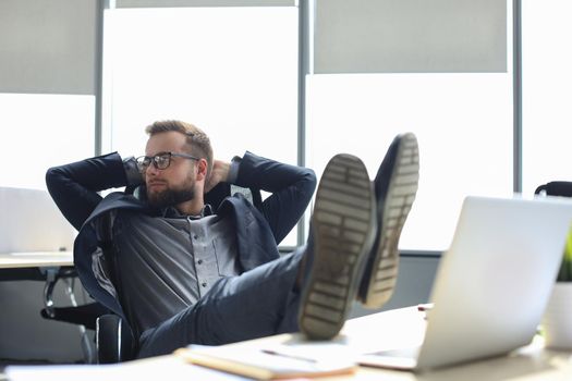 Young modern businessman keeping hands behind head and smiling while sitting in the office