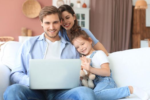 Positive friendly young parents with smiling little daughter sitting on sofa together answering video call on laptop and waving hand in greeting while relaxing at home on weekend