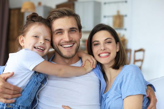 Positive friendly young parents with smiling little daughter sitting on sofa together while relaxing at home on weekend