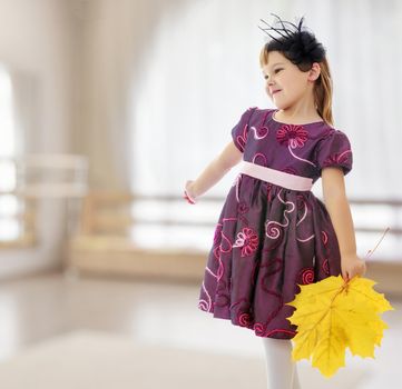 In a room with a large semi-circular window. Caucasian little girl dressed in brown dress. She is holding a bouquet of maple leaves. Turning sideways to the camera.
