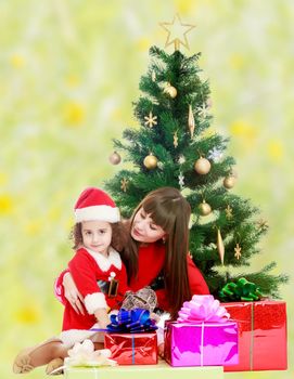 Beautiful mother and daughter , dressed in the costume of Santa Claus around the Christmas tree surrounded by heaps of gifts.Bright,floral yellow-green blurred background.