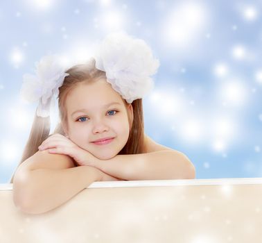 Gentle long-haired little girl with big white bows on the head . She leaned on the windowsill. Close-up.Blue Christmas background with white snowflakes.