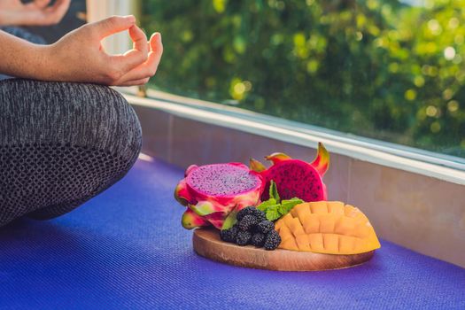 hand of a woman meditating in a yoga pose, sitting in lotus with fruits in front of her dragon fruit, mango and mulberry.