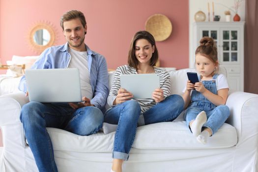 Father, mother and daughter using electronic devices sitting on sofa at living room