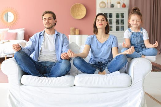 Calm young family with little daughter sit on couch practice yoga together, happy parents with small preschooler girl child rest on sofa meditate relieve negative emotions on weekend at home