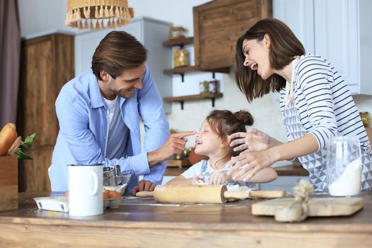 Cute little girl and her parents are having fun while cooking in kitchen at home together