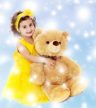 Joyful little girl in a yellow dress and bow on her head sitting on the floor. Girl hugging a big Teddy bear.Blue winter background with white snowflakes.