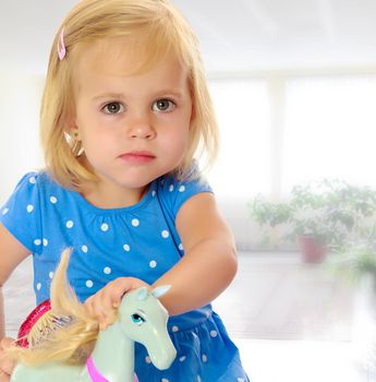 Cute little blonde girl playing with a toy horse. Girl wearing a blue dress with polka dots.Against the background of a child's room . The concept of the holiday and the New year.