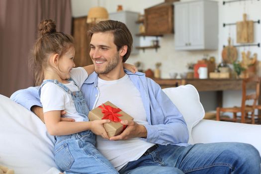 Cute kid daughter make surprise to daddy, little girl presents gift box to father sit on sofa. Father's day