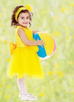 Laughing little girl in a bright yellow dress and bow on her head holding the ball. Girl posing sideways to the camera in full growth.Bright,floral yellow-green blurred background.