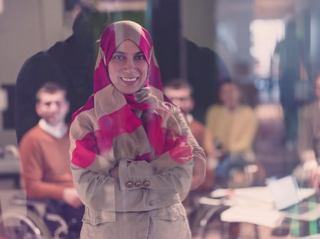 Portrait of Muslim woman wearing hijab in a modern open space coworking office. Middle eastern Successful Arab businesswoman in creative startup office. Disabled businessman in a wheelchair on team meeting in background.