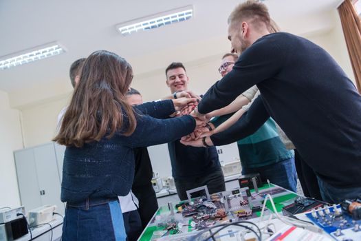 Group of young students in electronics classroom celebrating successfully finished project with holding their hands together, education and technology concept