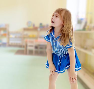 Cute little unkempt girl in a short blue dress. Girl looking to the side with his hands on his knees.In the rooms, with long shelves on which stand toys.