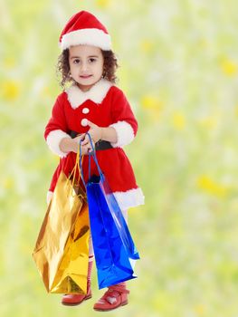Adorable little curly-haired girl in a coat and hat of Santa Claus,holding colorful shopping bags.Bright,floral yellow-green blurred background.