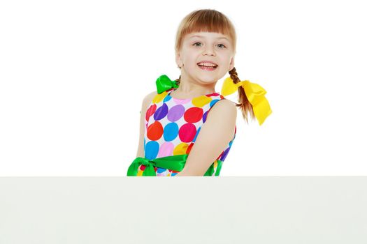 A beautiful Caucasian little blonde girl with long pigtails, in which large colored bows are braided, and a short bangs on her head. In a short summer dress.Looks because of the white obstacle.