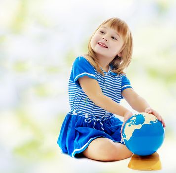 The concept of changing seasons in the life of a child. On the green and white blurred background. Nice little girl in a Sea blue dress sitting on the floor. Girl turns hand the globe.