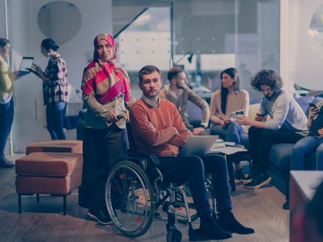 Disabled businessman in a wheelchair at work in modern open space coworking office on team meeting or brainstorming. Muslim businesswoman with hijab is assisting and helping on the project. Effective teamwork of volunteers concept in a startup business. A pregnant female coworker in the background. 