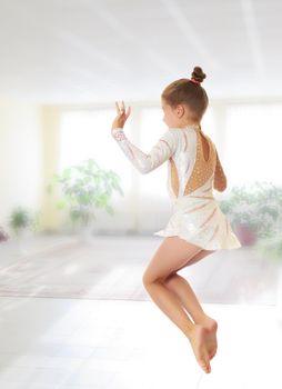 Beautiful little girl gymnast dressed in sports swimsuit, jumps high.On the background of the school hall with large Windows.