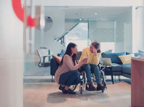 Sad and depressed disabled businesswoman in a wheelchair at the office getting support and assistance of female coworker friend. Modern open space office or hospital