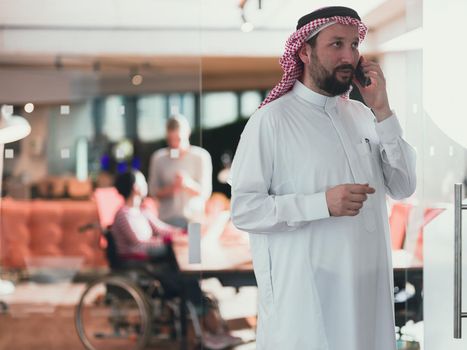 Middle eastern Arab businessman in the office meeting room speaking on the smartphone. Disabled businesswoman in wheelchair working together with the team in the background. High quality photo. Follow focus. 