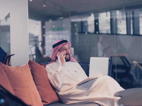 Mobile business communication and entrepreneurship. Arab businessman talking on smartphone and using a laptop computer, alone at open space coworking workplace or home office in the night. 