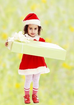 Cute little girl in a coat and hat of Santa Claus, holding a big green box , tied with a bow.Bright,floral yellow-green blurred background.