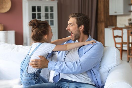 Smiling young father lying on couch at living room and play with happy little daughter