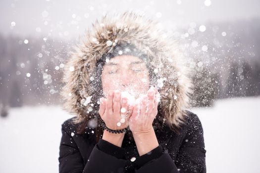 Happy young woman wearing winter clothes while blowing snow on snowy day with snowflakes around her in beautiful winter forest