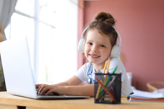 Smiling little girl in headphones handwrite study online using laptop at home, cute happy small child in earphones take Internet web lesson or class on PC