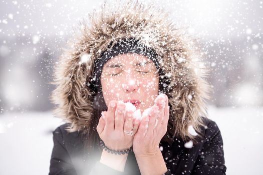 Happy young woman wearing winter clothes while blowing snow on snowy day with snowflakes around her in beautiful winter forest