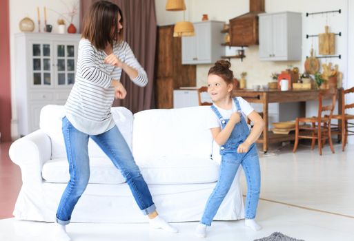 Cheerful mother with little daughter dancing at favourite song in living room at home