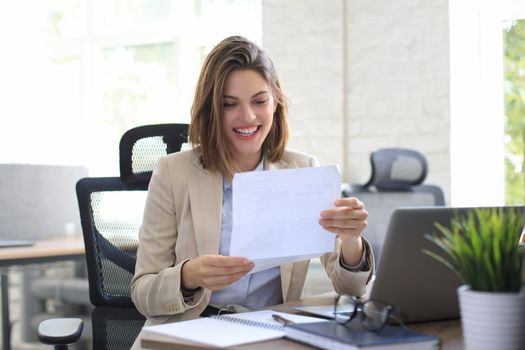 Attractive cheerful business woman checking paper documents in office, working on laptop