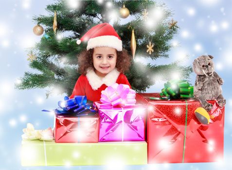 The beautiful, curly little girl in a suit and hat of Santa Claus sits near Christmas tree with bunch of gifts.Blue winter background with white snowflakes.