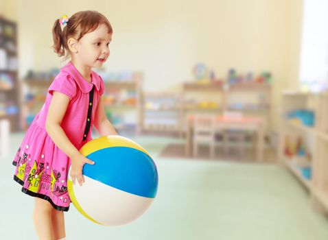 Beautiful little girl in a pink dress throwing a big striped ball. Turned sideways. Close-up.On blurred background the great hall of the kindergarten, with long racks where there are toys.