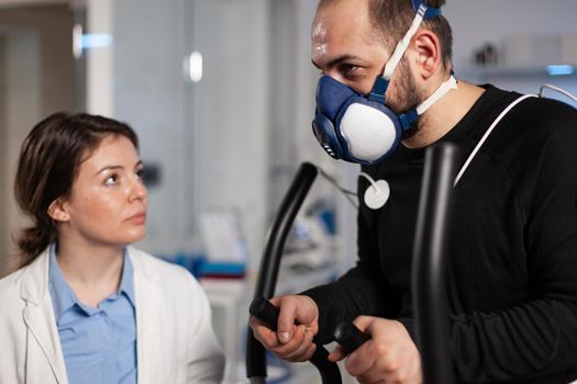 Sportman with mask and electrodes sensor on it doing fitness exercise working at body endurance during medical workout in clinical laboratory. Physician doctor monitoring EKG data using hi-tech