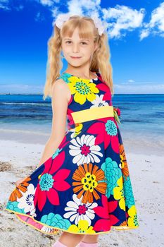Beautiful little girl with long blonde ponytails on the head and white bows in bright summer dress keeps the dress over the edge.On the background of sea beach, warm sea and blue sky with clouds.