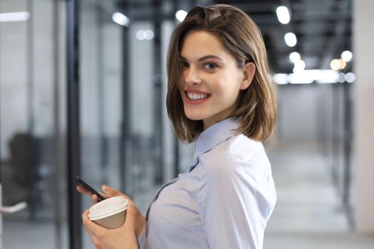 Businesswoman walking along the office corridor with paper cup and smartphone