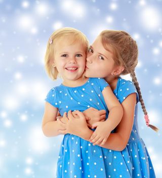 Two charming little girls, sisters , in identical blue dresses with polka dots. Older sister kissing the younger on the cheek.On a blue background with large, white,