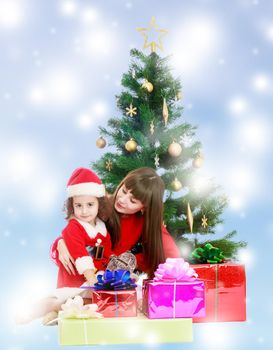 Beautiful mother and daughter , dressed in the costume of Santa Claus around the Christmas tree surrounded by heaps of gifts.Blue winter background with white snowflakes.