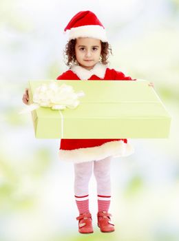 Cute little girl in a coat and hat of Santa Claus, holding a big green box , tied with a bow.white-green blurred abstract background with snowflakes.