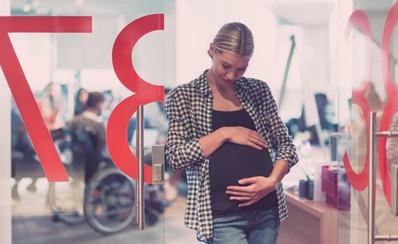 Portrait of pregnant businesswoman gently holding belly in a modern office team meeting of coworkers in background. The disabled coworker in a wheelchair 