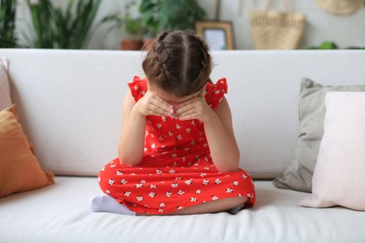 Sad little girl sitting on sofa alone at home