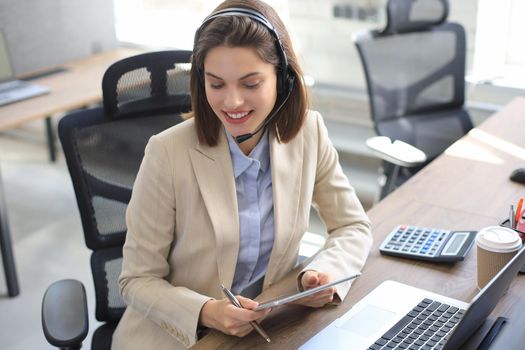 Cheerful female manager sitting at office desk and performing corporate tasks using wireless connection on digital gadgets