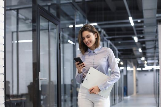 Businesswoman walking along the office corridor with documents and using smartphone