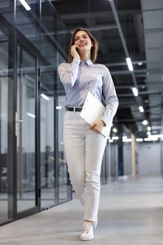 Businesswoman walking along the office corridor with documents and using smartphone