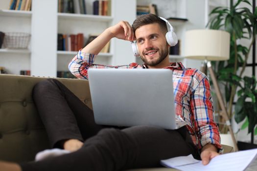 Concentrated young freelancer businessman sitting on sofa with laptop, working remotely online at home