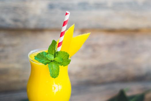 Juicy smoothie from mango in glass with striped red straw and with a mint leaf on old wooden background. Healthy life concept, copy space.