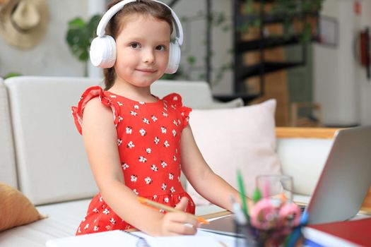 Smiling little girl in headphones handwrite study online using laptop at home, cute happy small child in earphones take Internet web lesson or class on PC